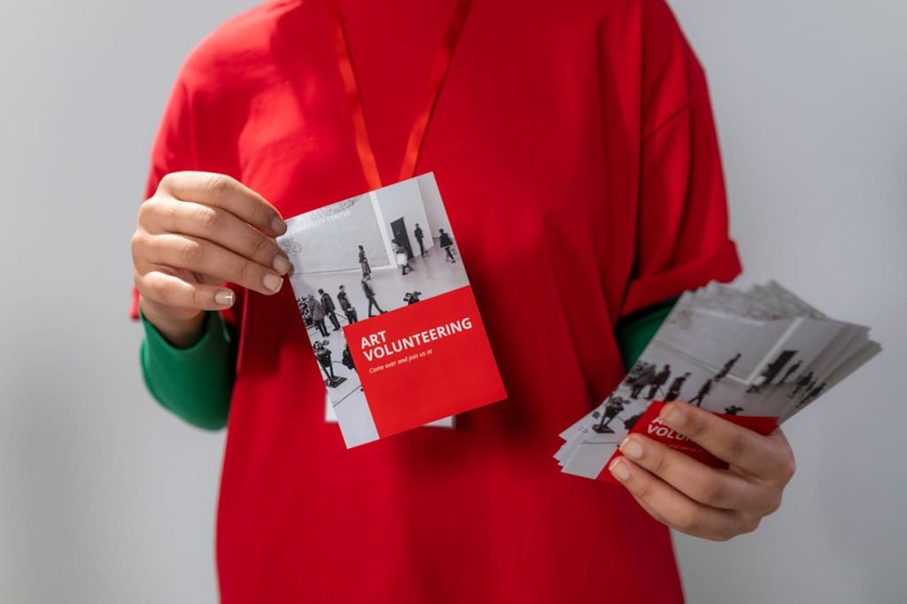 close-up-photo-of-red-brochure-6565749 Close-up of a volunteer holding art volunteering flyers highlighting community service.